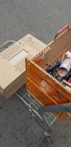 Overhead-Aufnahme: Einkaufskorb mit Baumarktmaterialien auf grauem Asphalt — Kampagnenmotiv für Handwerk, Bau, DIY-Indust…
