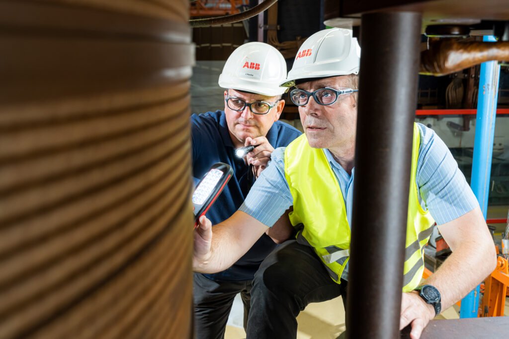 Endabnahme-Reportage ABB Industrie — zwei Techniker inspizieren Komponente vor Ort, Studio-dokumentarisch für B2B-Maschinenb…